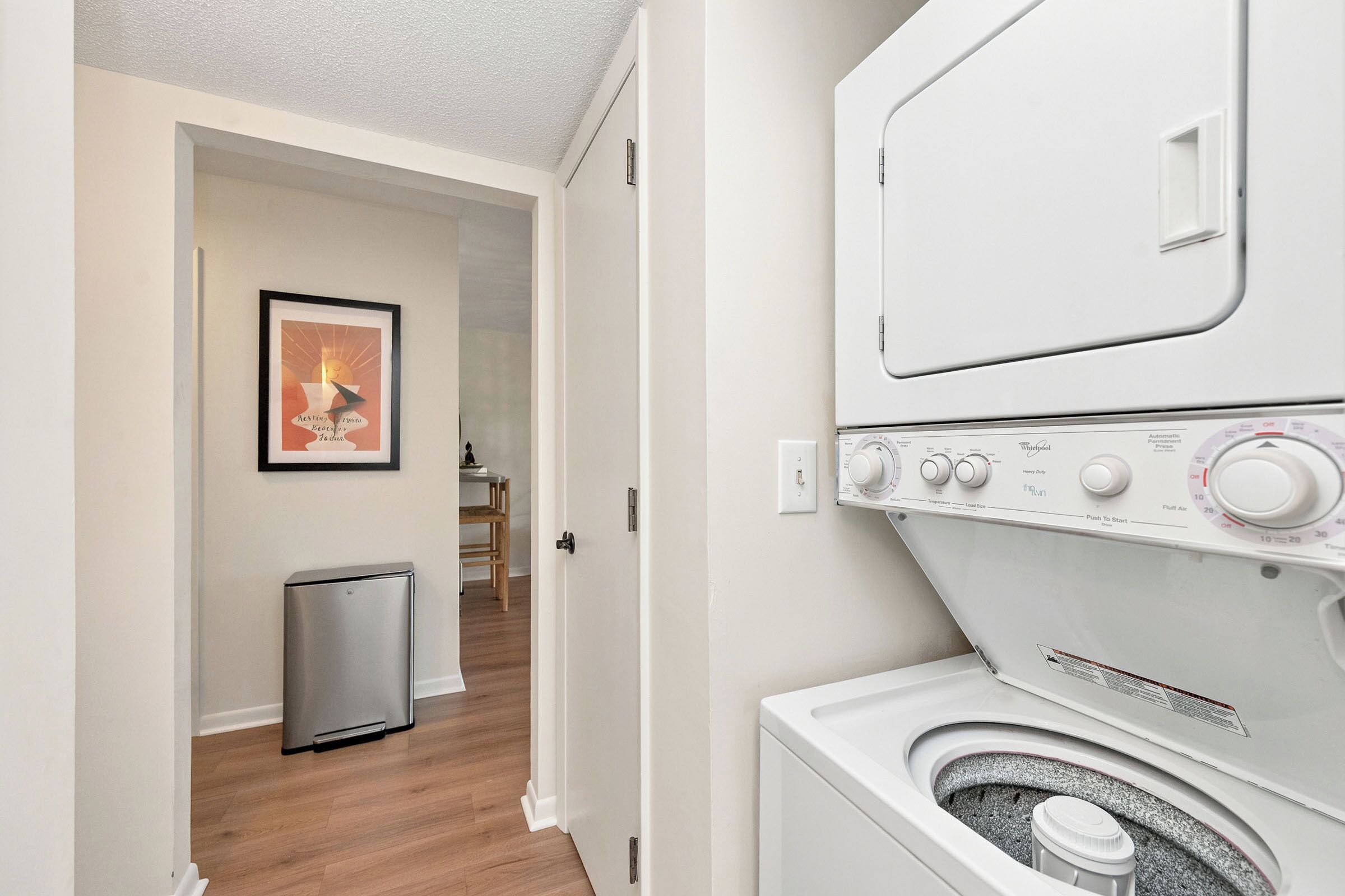 A laundry room with stacked washers and dryers, a small trash can, and a framed picture on the wall.
