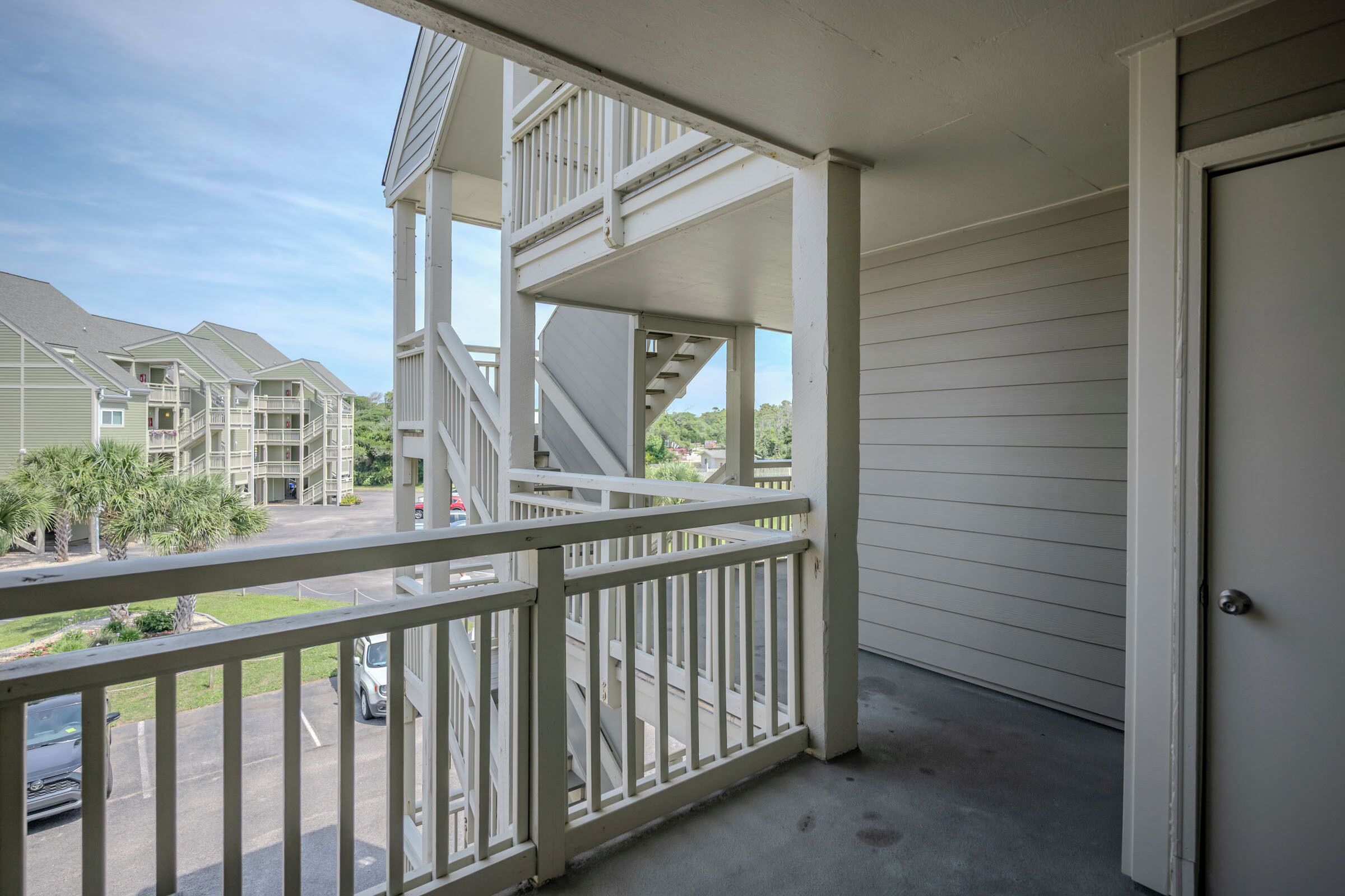 A balcony with a white railing, a door to the right, and a view of a parking lot and buildings in the background.