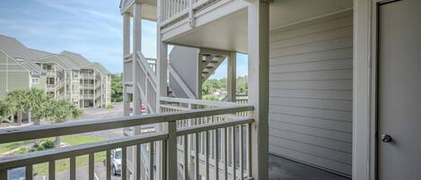 A balcony with a white railing, a door to the right, and a view of a parking lot and buildings in the background.