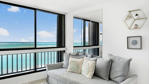A modern living room with a large window overlooking a beach, a grey sofa with decorative pillows, and a geometric wall decoration.
