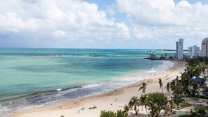 A beach with clear turquoise waters, a sandy shore, and a row of buildings in the background.