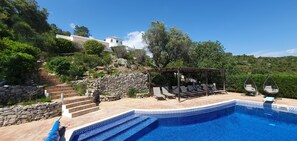 A swimming pool with a stone wall and steps leading up to it, surrounded by greenery and a pergola with chairs. - Casa Estrela, Luxury Algarve Villa, Stunning Views, Private Pool, Gardens, Fibre (Santa Bárbara de Nexe)