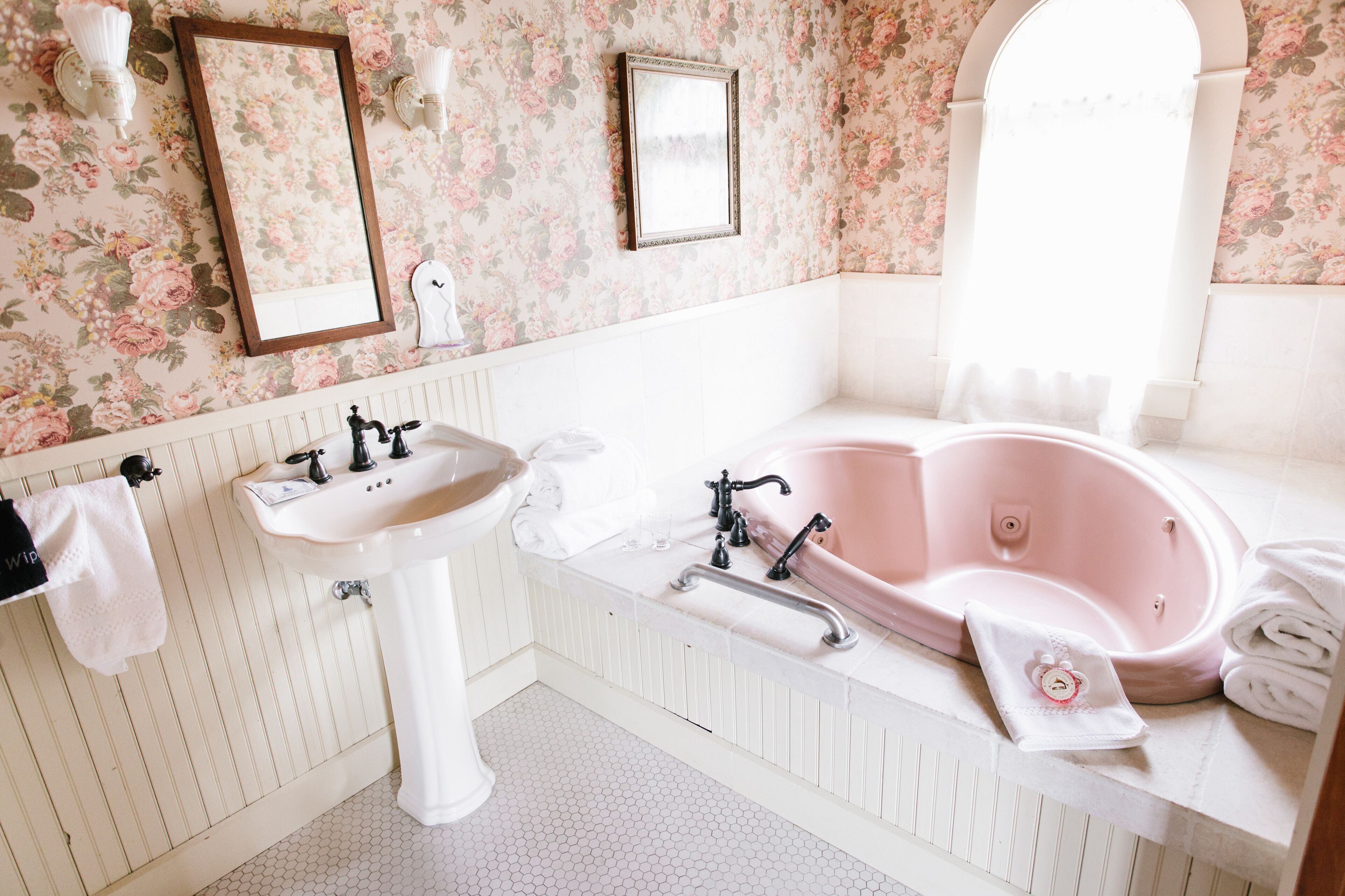 A bathroom with a white sink, a pink bathtub, floral wallpaper, and a window.