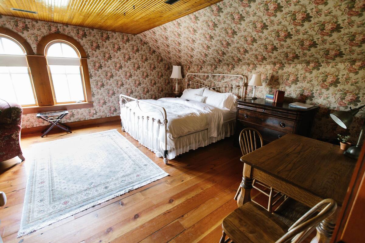 A bedroom with a wooden ceiling, floral wallpaper, a bed with white linens, a wooden dresser, and a small table with a lamp.