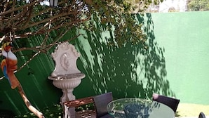 Un patio al aire libre con una mesa redonda de vidrio y sillas de mimbre, rodeado por un muro verde y una urna decorativa.