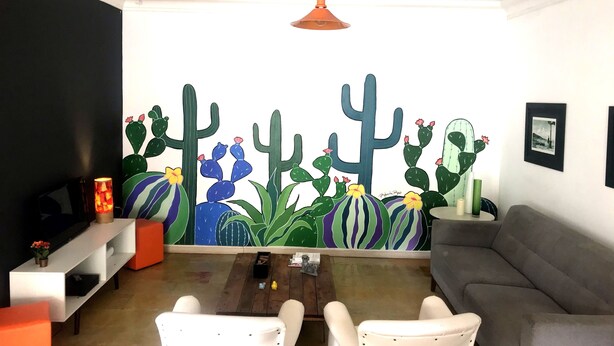 Una sala de estar con un gran mural de cactus, un sofá gris, sillones blancos y una mesa de centro de madera.