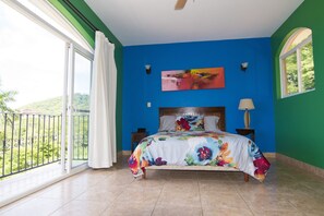 A bedroom with a large bed, a view of greenery through a balcony, and a painting on the wall.