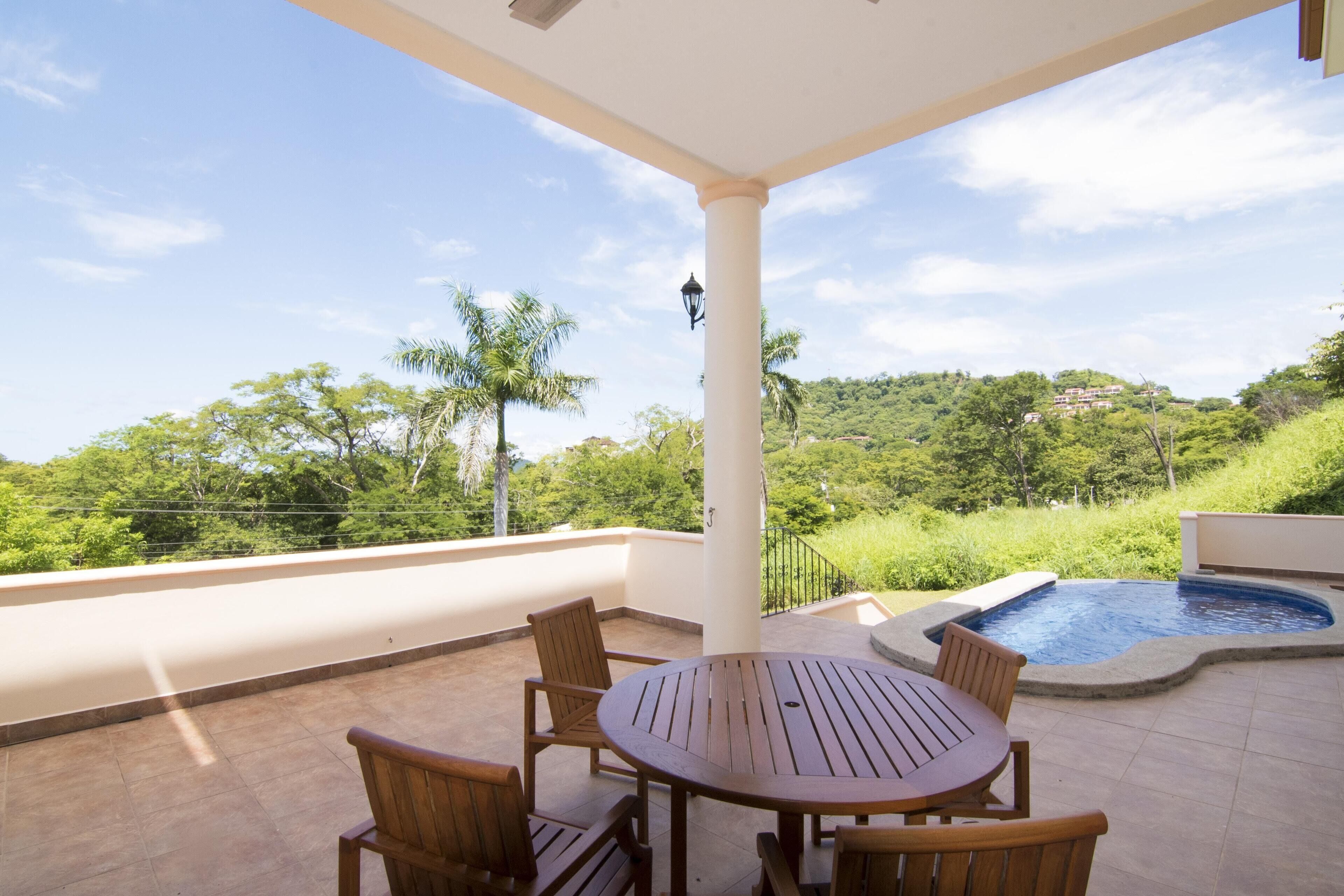 A covered outdoor area with a round wooden table and chairs, a small pool, and a view of a lush green landscape.