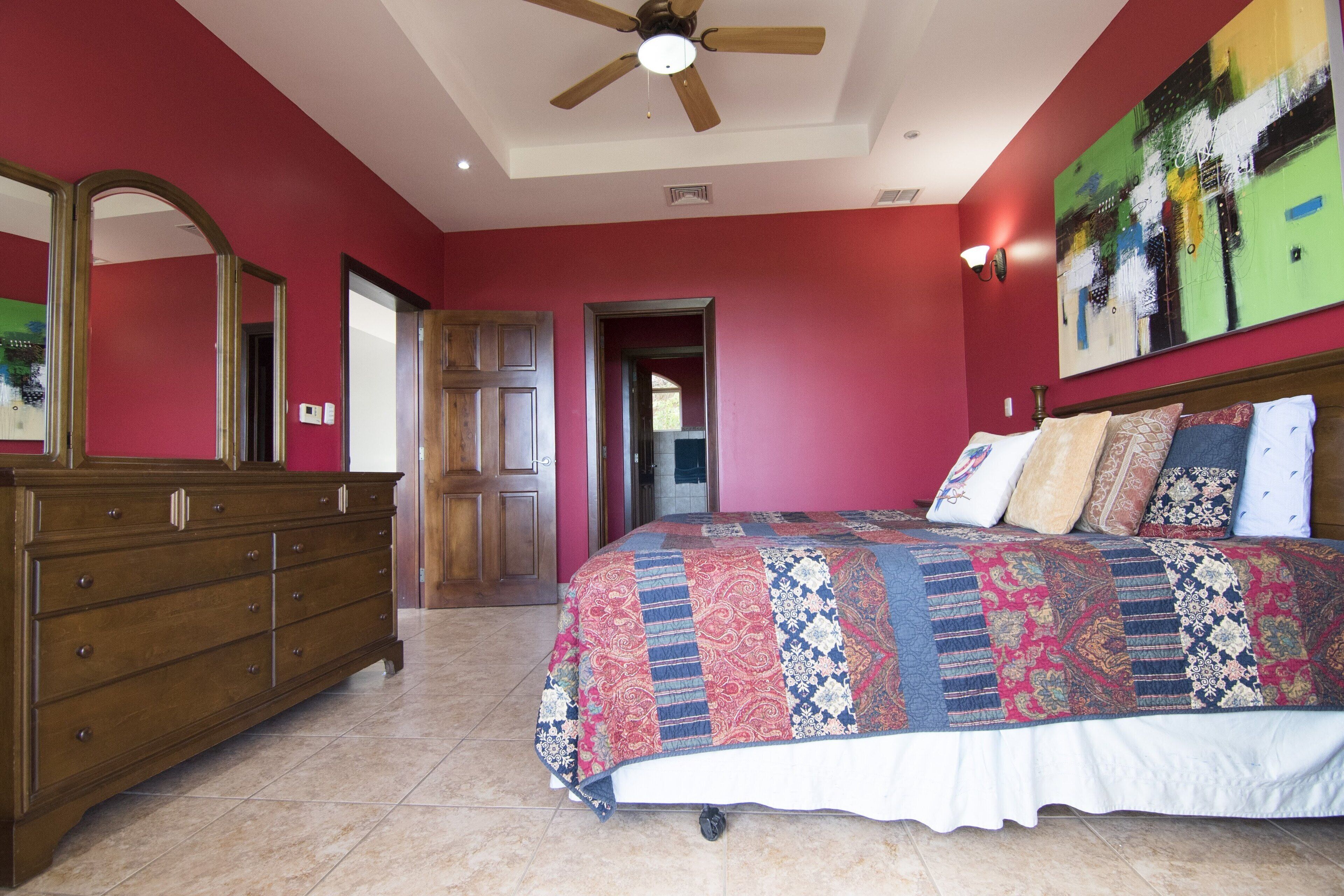 A bedroom with a large bed, a wooden dresser, a ceiling fan, and a painting on the wall.