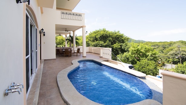 Une piscine à bordures incurvées, un patio couvert aménagé avec du mobilier d’extérieur et une vue sur un paysage verdoyant luxuriant.