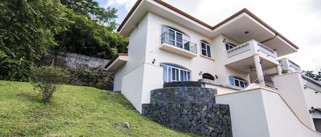 A two-story house with a stone retaining wall, a balcony, and a small tree.
