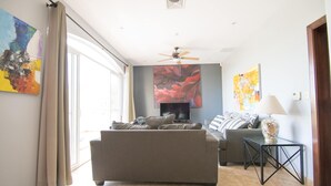 Un salon avec un canapé, une table basse et un téléviseur. Il y a des rideaux à la fenêtre et des œuvres d’art accrochées aux murs.