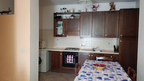 A compact kitchen with wooden cabinets, a built-in washing machine, and a dining table with a fish-patterned tablecloth. - App.in semi-detached villa private garden and parking shared pool (Imperia)