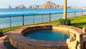 A hot tub with a stone border, a metal fence, and a view of the sea and mountains in the background.
