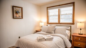 A neatly made bed with a patterned bedspread, two bedside lamps, a nightstand, and a framed picture on the wall. - Brook Road Condo (Goshen)