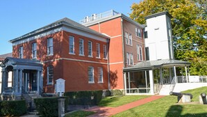 A red brick building with a classical facade, a covered walkway, and a well-maintained lawn. - Brook Road Condo (Goshen)