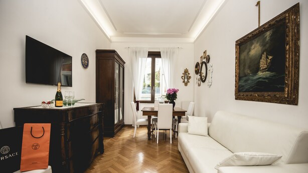 A modern living room with a white sofa, a wooden dining table with chairs, a TV mounted on the wall, and a painting. - St. Peter's Heart -honeymoon luxury apartment Rome (roma)