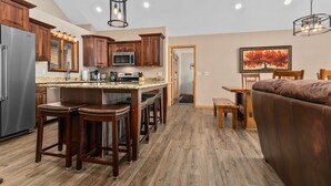A modern kitchen with wooden cabinets, a stainless steel refrigerator, and a granite countertop. There is a dining area with a brown leather couch and a painting of a tree. - Full Rec Lake w/pontoon to rent! 🏖 Beach. 🐶 friendly. (Wautoma)