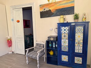 Una habitación con una puerta blanca, una silla, un armario azul con azulejos decorativos, un cuadro de un atardecer y una pequeña planta en maceta.