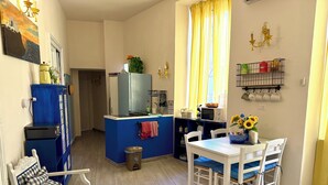 A compact kitchen with a blue refrigerator, microwave, and sink. There is a dining table with chairs and a small window with yellow curtains.