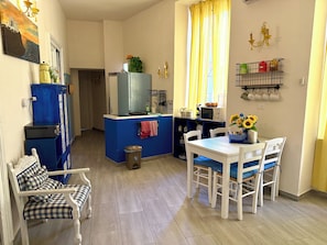 Una cocina compacta con un refrigerador azul, microondas y lavamanos. Hay una mesa de comedor con sillas y una pequeña ventana con cortinas amarillas.