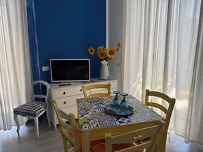 A dining area with a table set for four, a television on a white cabinet, and a vase with sunflowers on the wall.