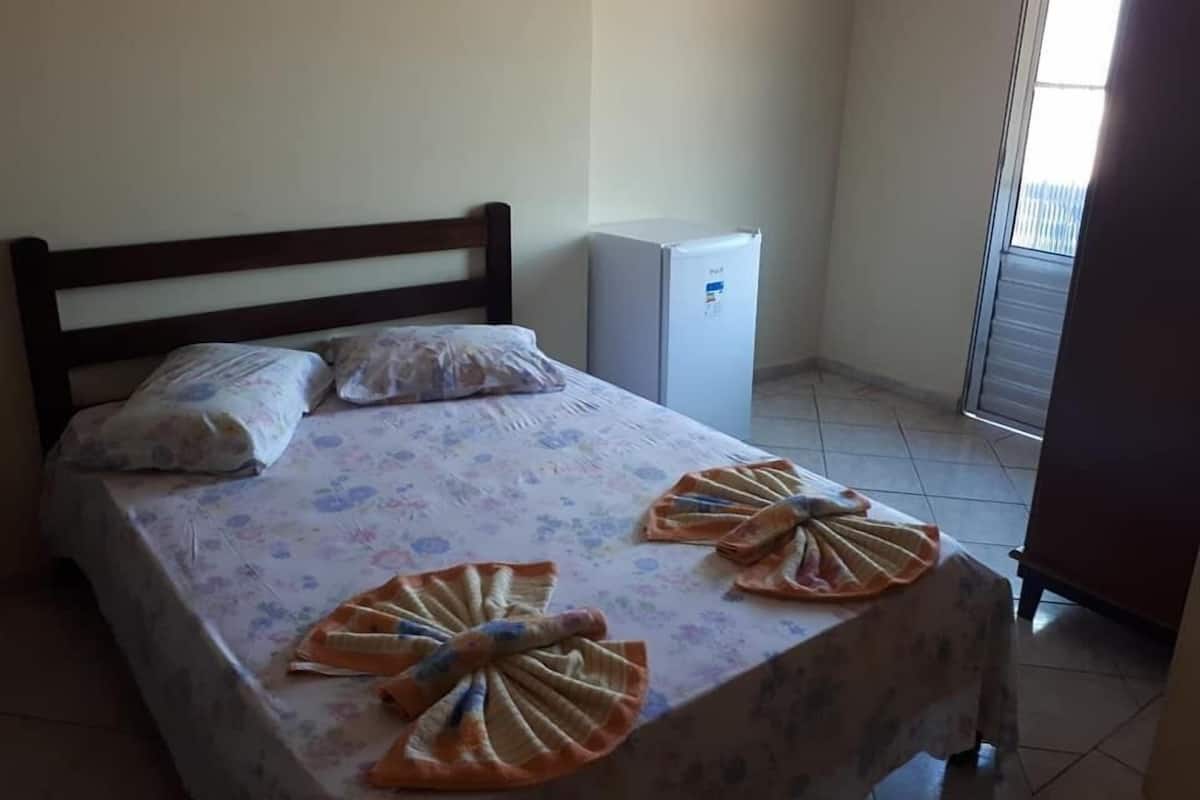 A bed with floral bedding, two pillows, and towels arranged in a fan shape. A small white refrigerator is against the wall.