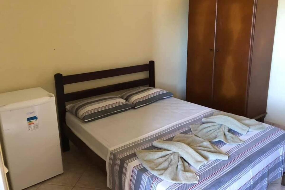 A single bed with a striped bedspread and pillows, a wooden wardrobe, and a small white appliance.