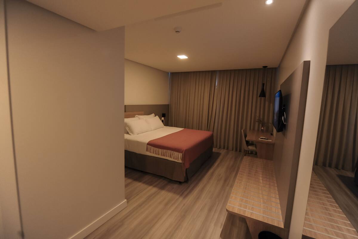 Una habitación de hotel con cama, escritorio, silla, televisor y cortina.