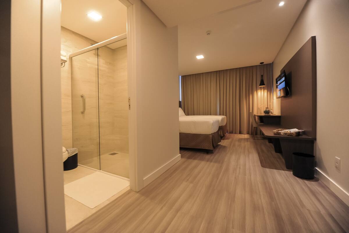 Una habitación de hotel moderna con ducha de vidrio, cama, televisor y un pequeño escritorio.