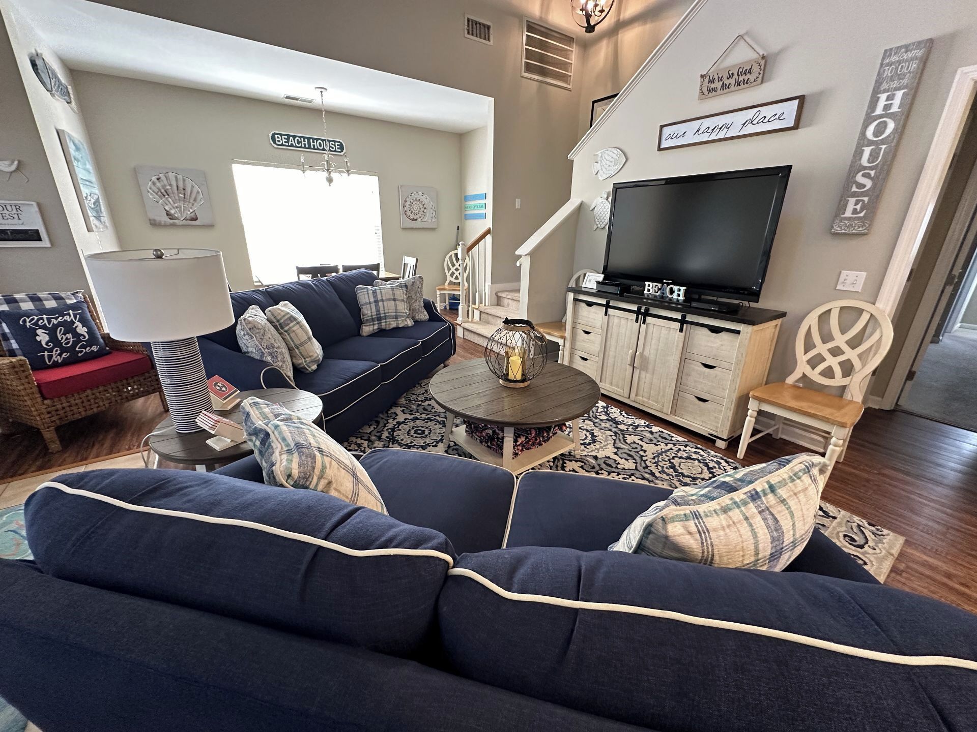 Uma sala de estar com um sofá azul escuro, uma televisão sobre um móvel de madeira e uma placa com a inscrição 'BEACH HOUSE'.