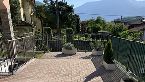 A paved terrace with brick flooring, bordered by metal fencing, and featuring planters with small trees. - Pool apartment in quiet and central area (Colico , Lombardei)