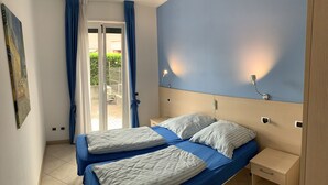 A room with two beds, a nightstand, a wall-mounted light, and a view through a glass door. - Pool apartment in quiet and central area (Colico , Lombardei)
