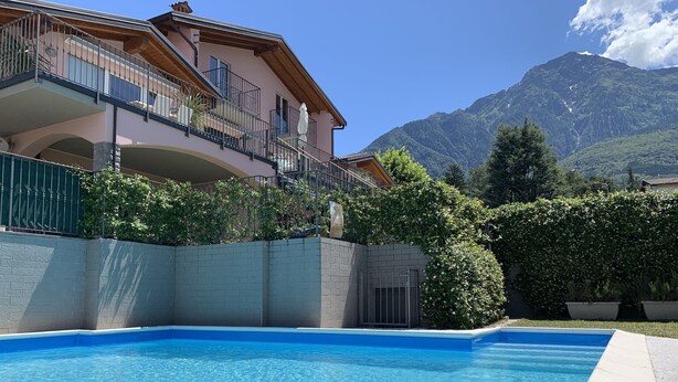 A swimming pool with a mountain backdrop, a multi-story building with balconies, and a clear sky. - Pool apartment in quiet and central area (Colico , Lombardei)