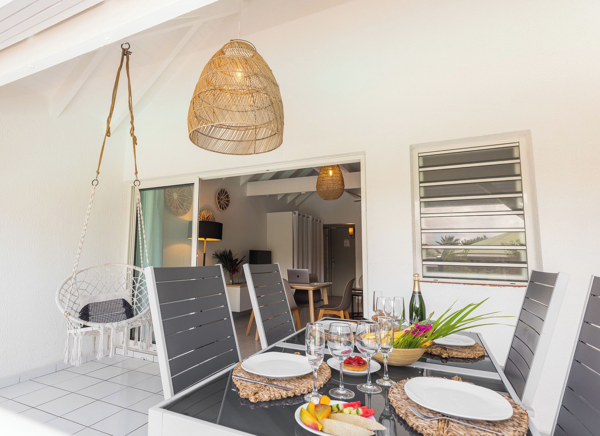 Un comedor con una mesa preparada para cuatro, incluyendo platos, copas y una botella de champán. Encima de la mesa hay una lámpara colgante de mimbre, y una silla blanca con asiento trenzado. La habitación tiene un diseño moderno, con un ventanal grande y una puerta que conduce a otra área.