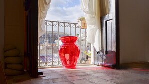 Ein roter Vasen auf einem Balkon mit Metallgeländer, einer Tür und Blick auf Gebäude im Hintergrund.