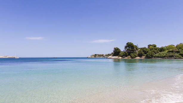 Ein Strand mit klarem türkisblauem Wasser, sandigem Ufer und einer kleinen Insel mit Bäumen und Gebäuden.