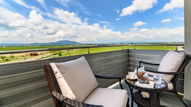 Un balcon donnant sur un champ verdoyant et des montagnes, avec des fauteuils en osier et une petite table où sont disposées des boissons.
