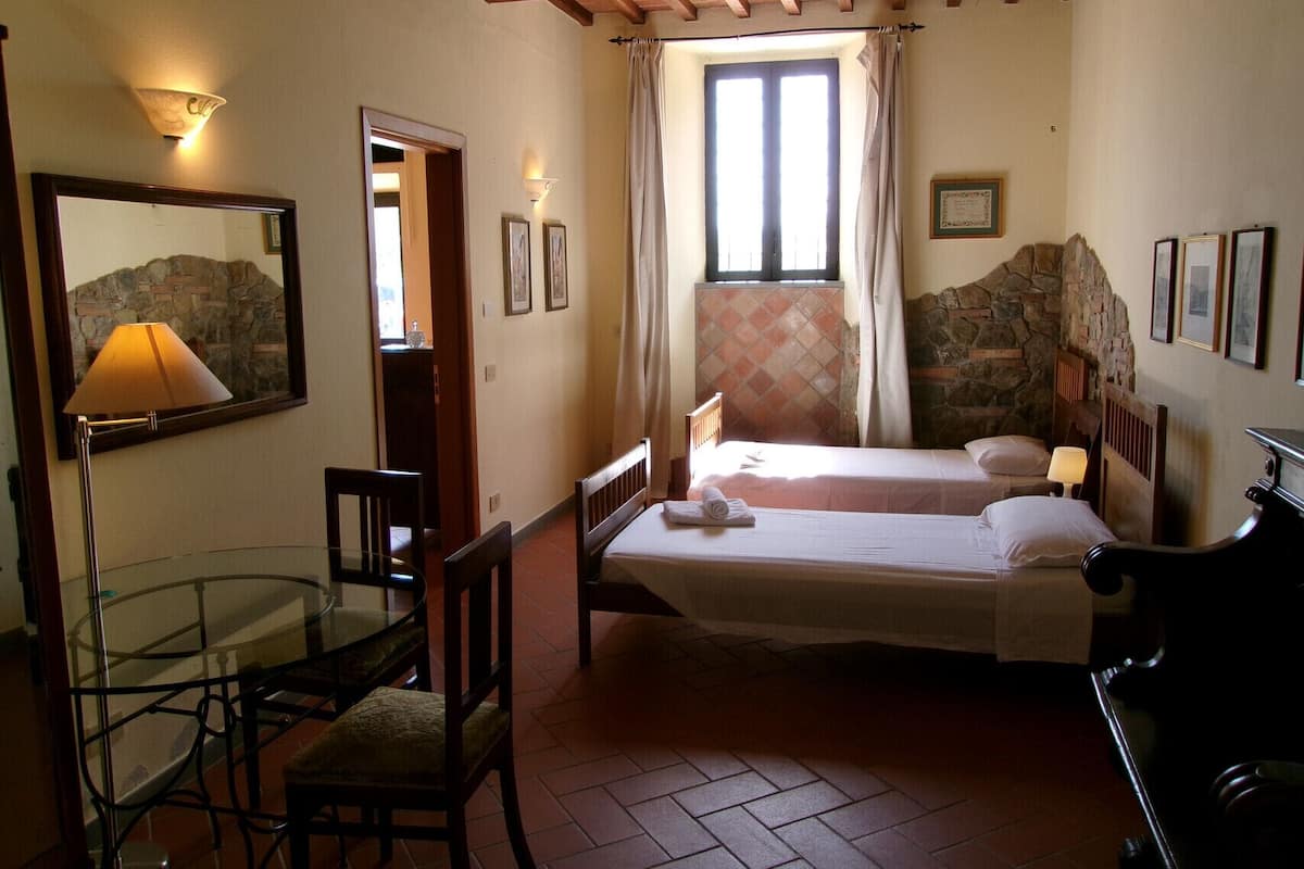 A bedroom with two beds, a desk, a chair, a lamp, and a stone wall.