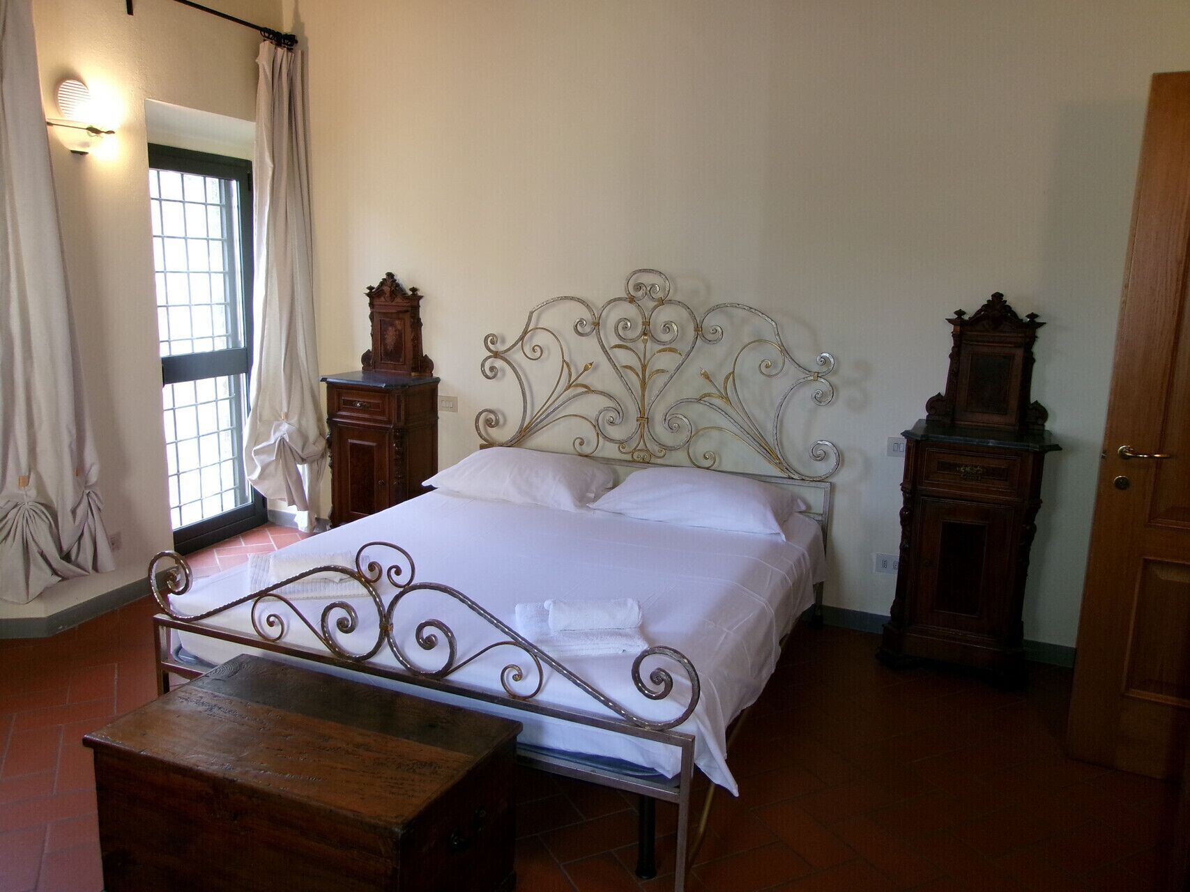 Een slaapkamer met een metalen bedframe, een houten dressoir, een kleine tafel en een raam met gordijnen.