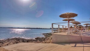 A beachside area with lounge chairs and umbrellas, a concrete walkway, and a rocky shoreline.
