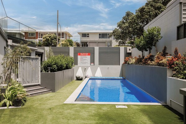 A rectangular pool with a clear blue surface, surrounded by a low white fence, and a sign indicating pool area hours. - Sunny & Spacious, Downtown Mount, Beach & Pool (Mount Maunganui)