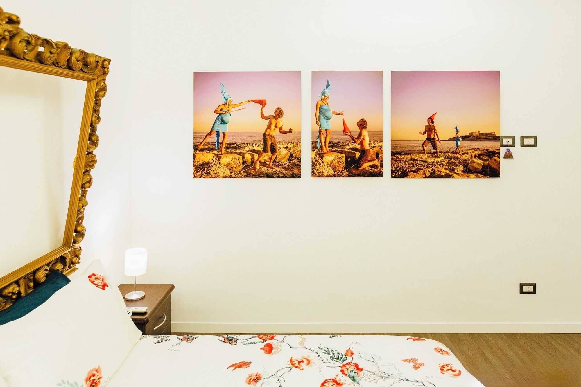 Un dormitorio con una cama, una mesita de noche, un espejo montado en la pared y tres cuadros con escenas de playa.