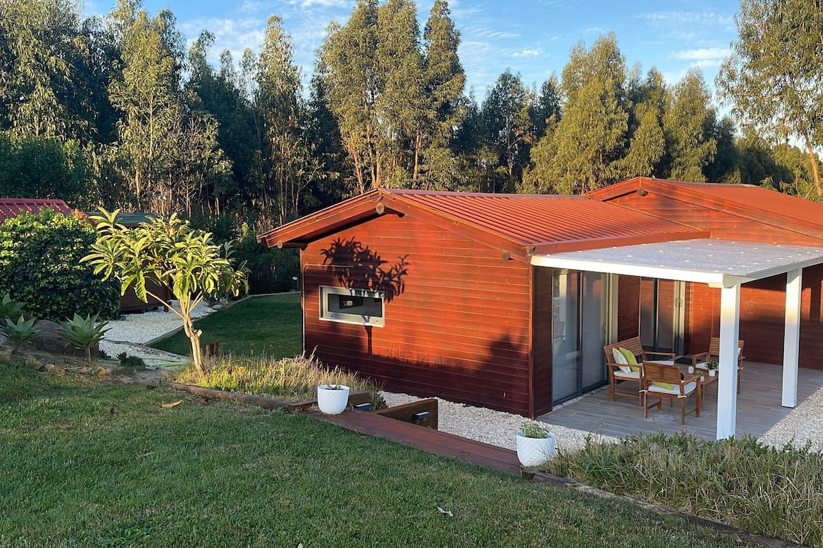 Een moderne houten blokhut met een overdekt terras, omringd door een verzorgde tuin en hoge bomen.