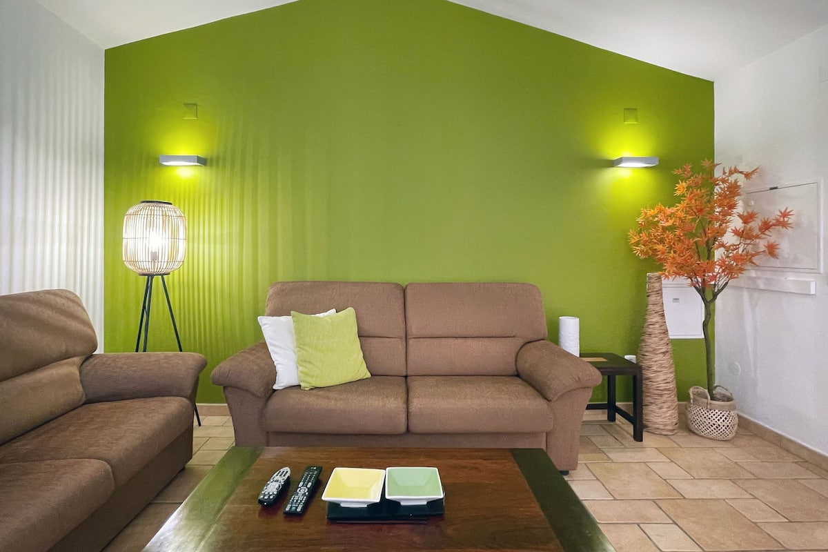 Een woonkamer met een groene accentmuur, een bruine bank, een salontafel met afstandsbedieningen en een bijzettafeltje met een plant.
