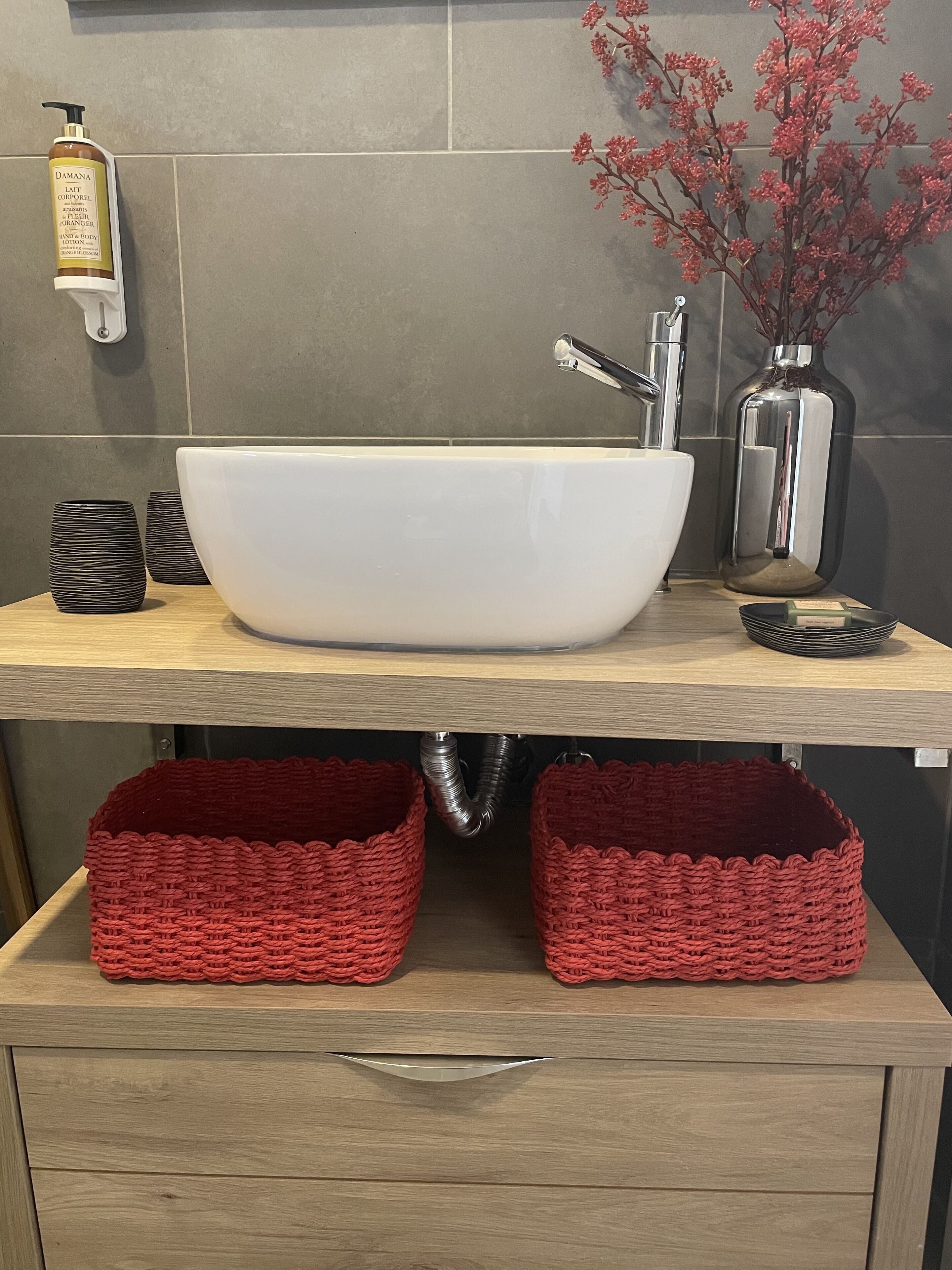 Un baño moderno con un lavabo blanco de diseño, un espejo, un florero rojo con flores y dos cestas rojas tejidas sobre un mueble de tocador de madera.