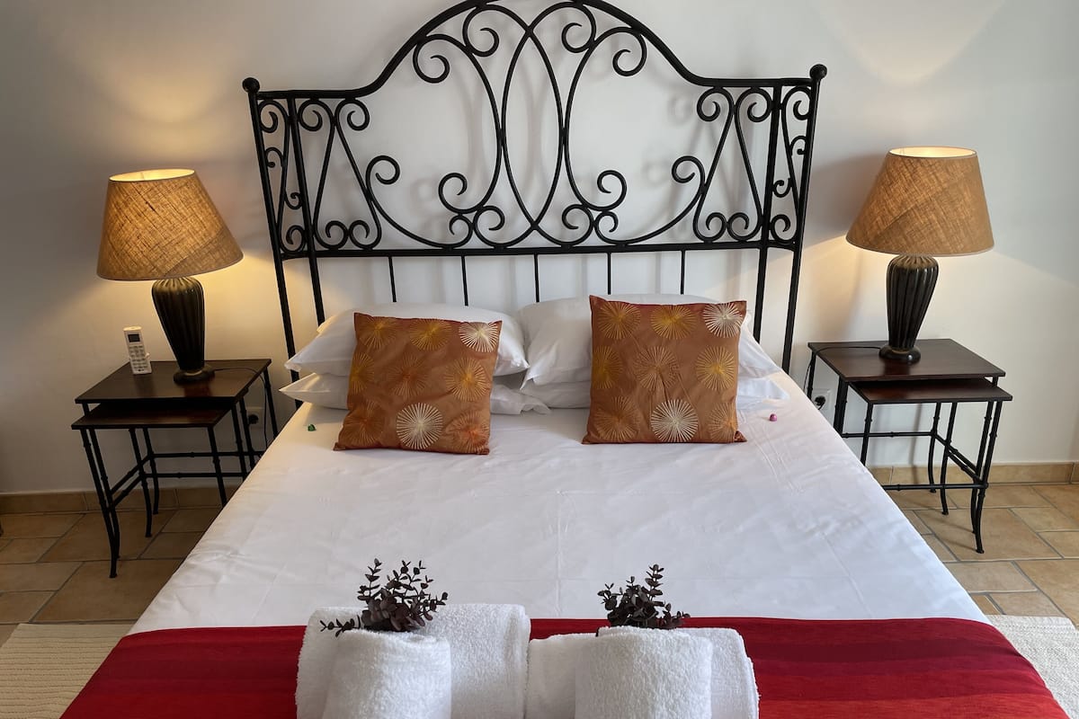 Een net opgemaakt bed met witte lakens en twee decoratieve kussens, met aan weerszijden nachtkastjes met lampen en een klein plantje.