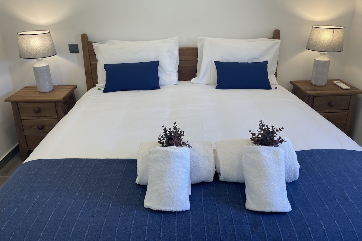 Een net opgemaakt bed met wit- en blauwe beddengoed, twee nachtkastjes met lampen, en een blauw gestreept vloerkleed.