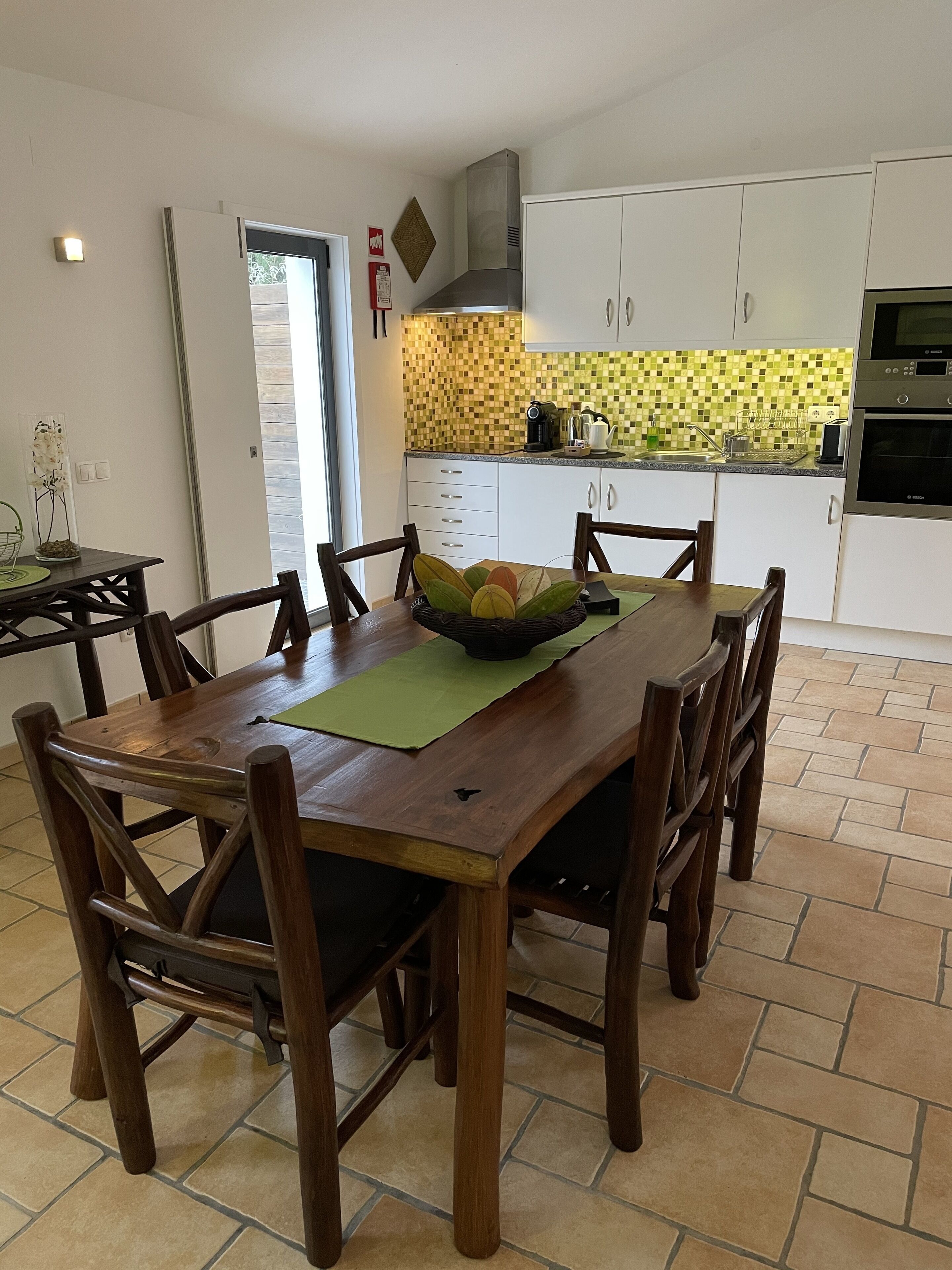 Un coin repas avec une table et des chaises en bois, une cuisine en arrière-plan avec des meubles blancs et un mur recouvert de carreaux, et une porte donnant sur l’extérieur.