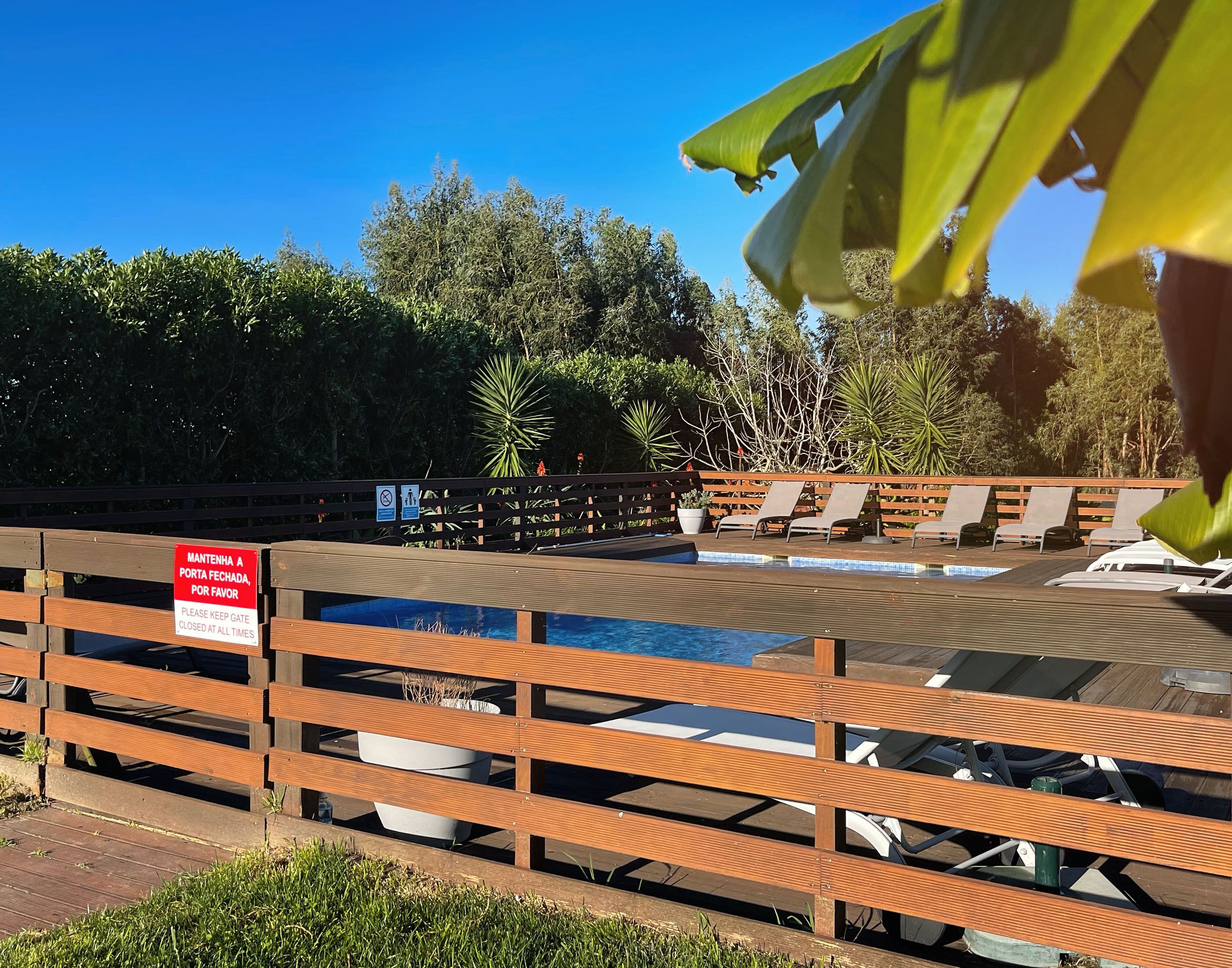 A pool area with wooden benches and lounge chairs, a closed gate sign, and lush greenery in the background.